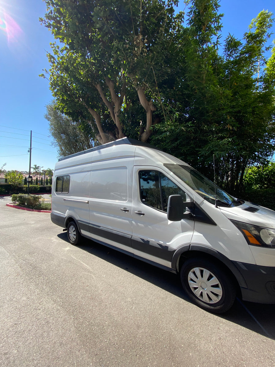 White Ford Transit RV rental van parked on sunny asphalt beside green trees.