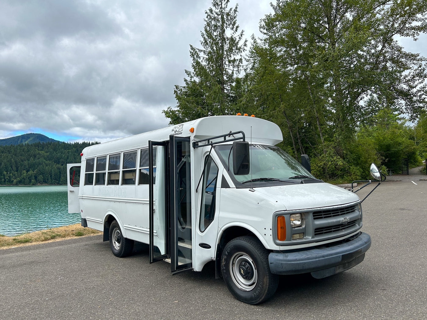 White bus-style rv rental parked at a scenic lake, nestled by mountains and forests, ideal for outdoor trips. — photo 2