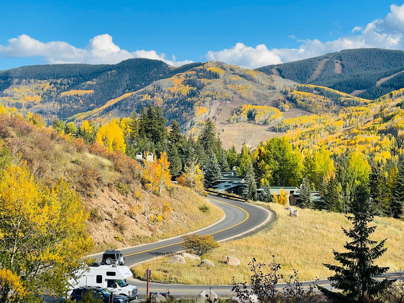 An RV rental navigates a winding mountain road, surrounded by vibrant fall foliage. Distant peaks reveal ski trails under a blue sky.
