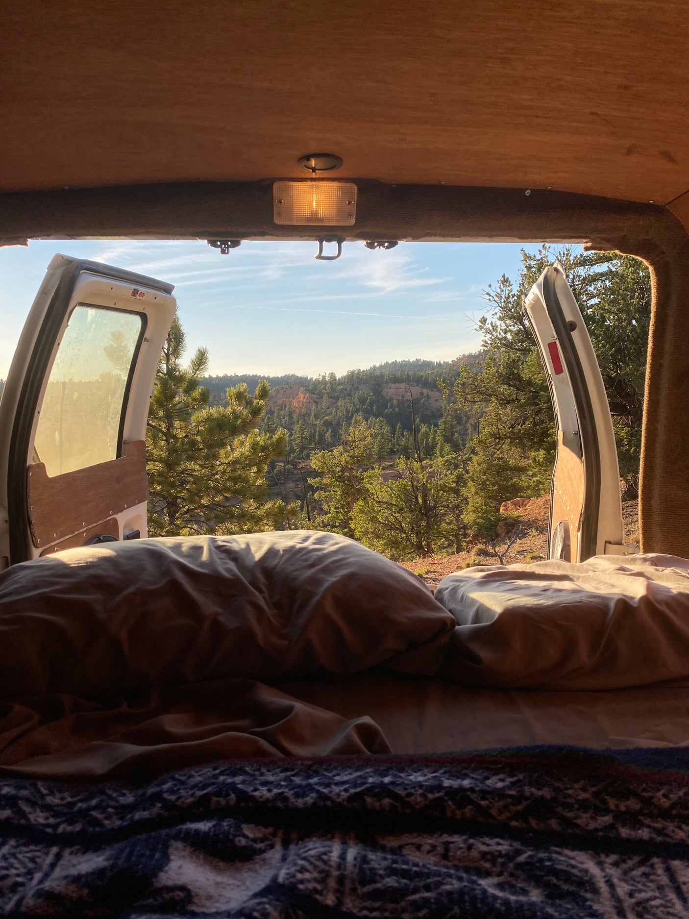Inside a cozy **RV rental**, a bed faces open rear doors revealing a serene mountain forest view, perfect for **van life** adventures.