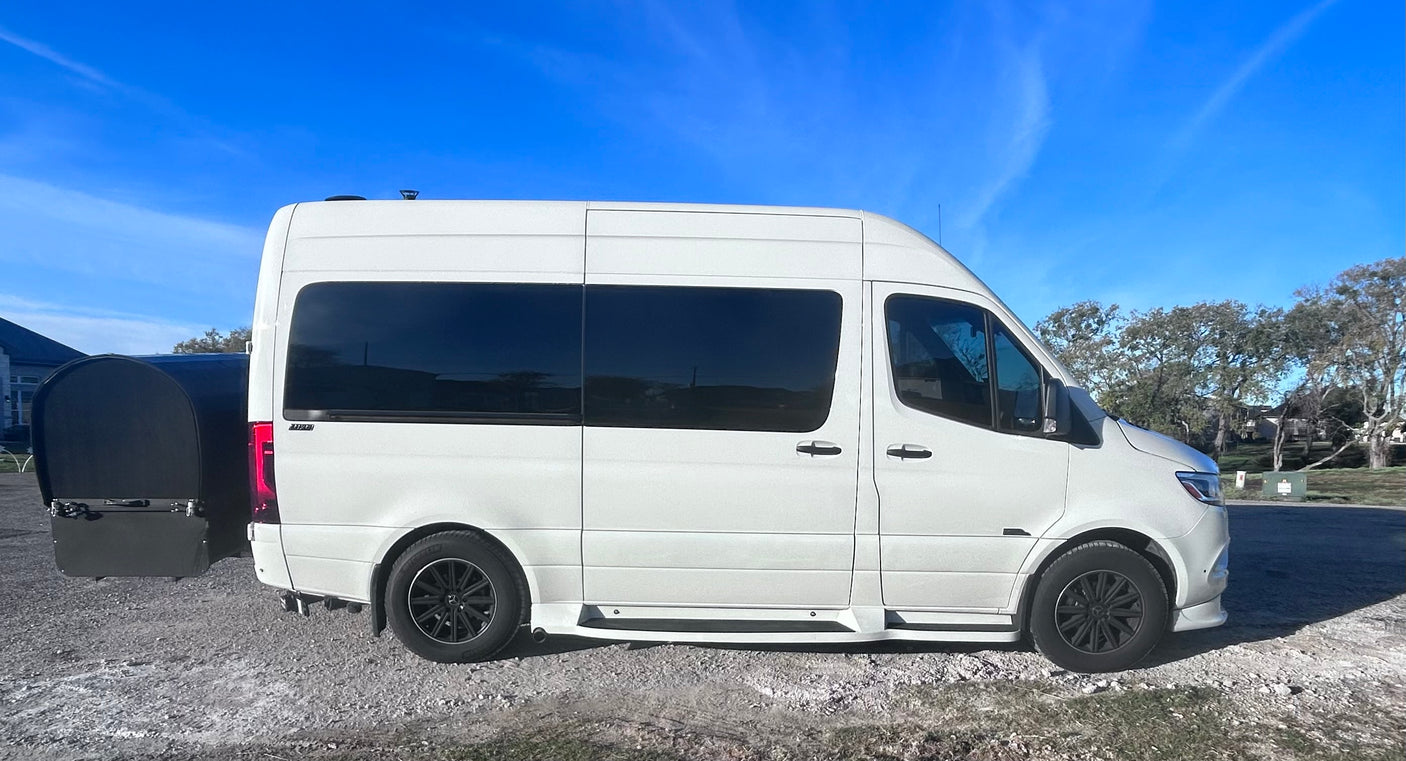 White Sprinter van, commonly used for RV rental, with a rear cargo carrier, parked on gravel under a blue sky.
