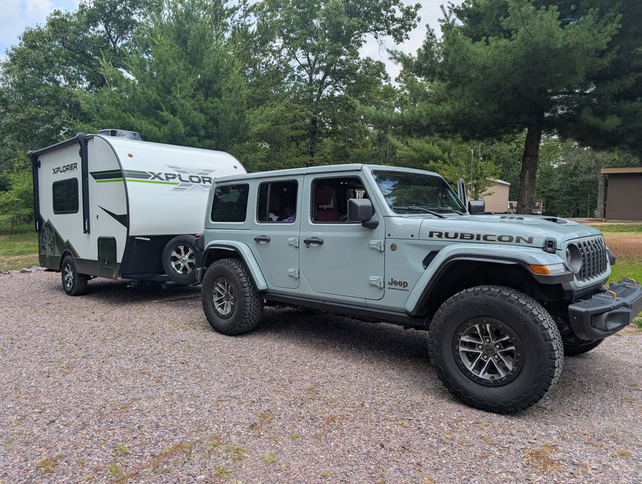 Light gray Jeep Rubicon towing an Xplorer rv rental trailer on a gravel lot, ready for outdoor adventure.