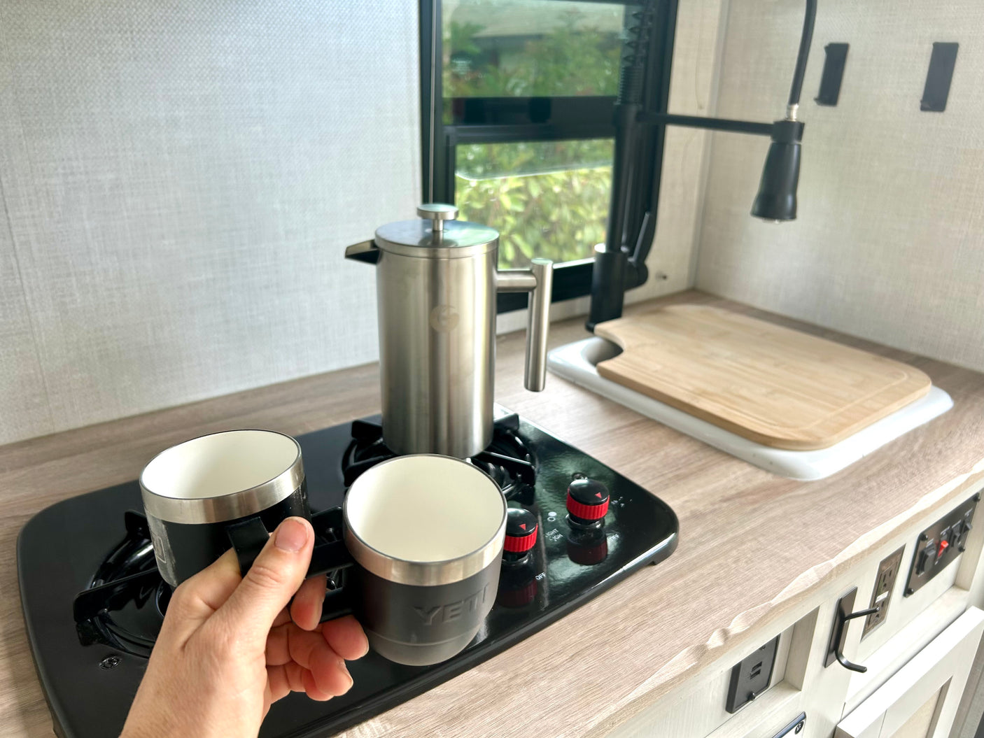 Enjoying coffee in an RV rental: a person holds a black Yeti mug next to a French press and another mug on a stovetop.