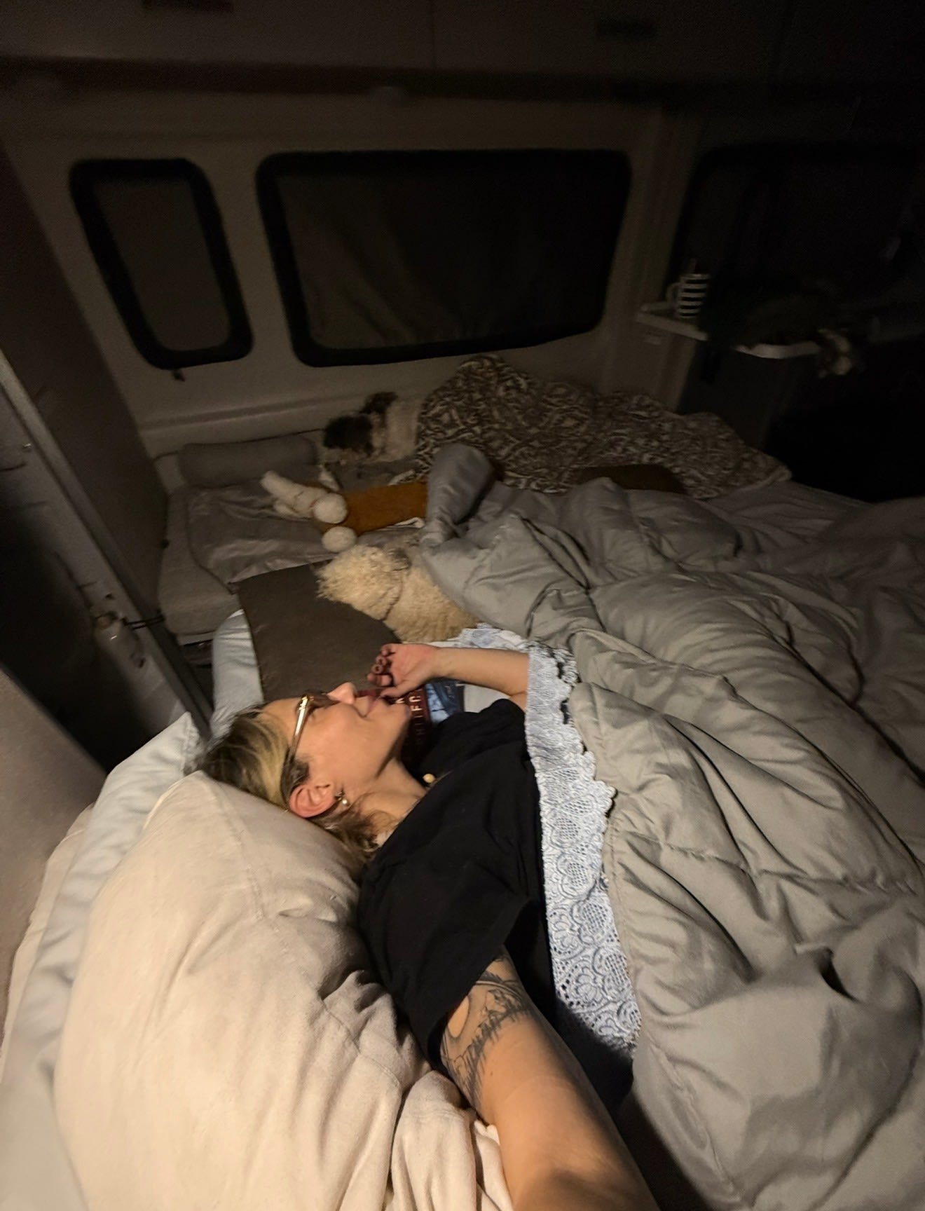 Cozy comfort in an RV rental. Person with glasses resting in bed under a comforter, a book and pets nearby in the camper.