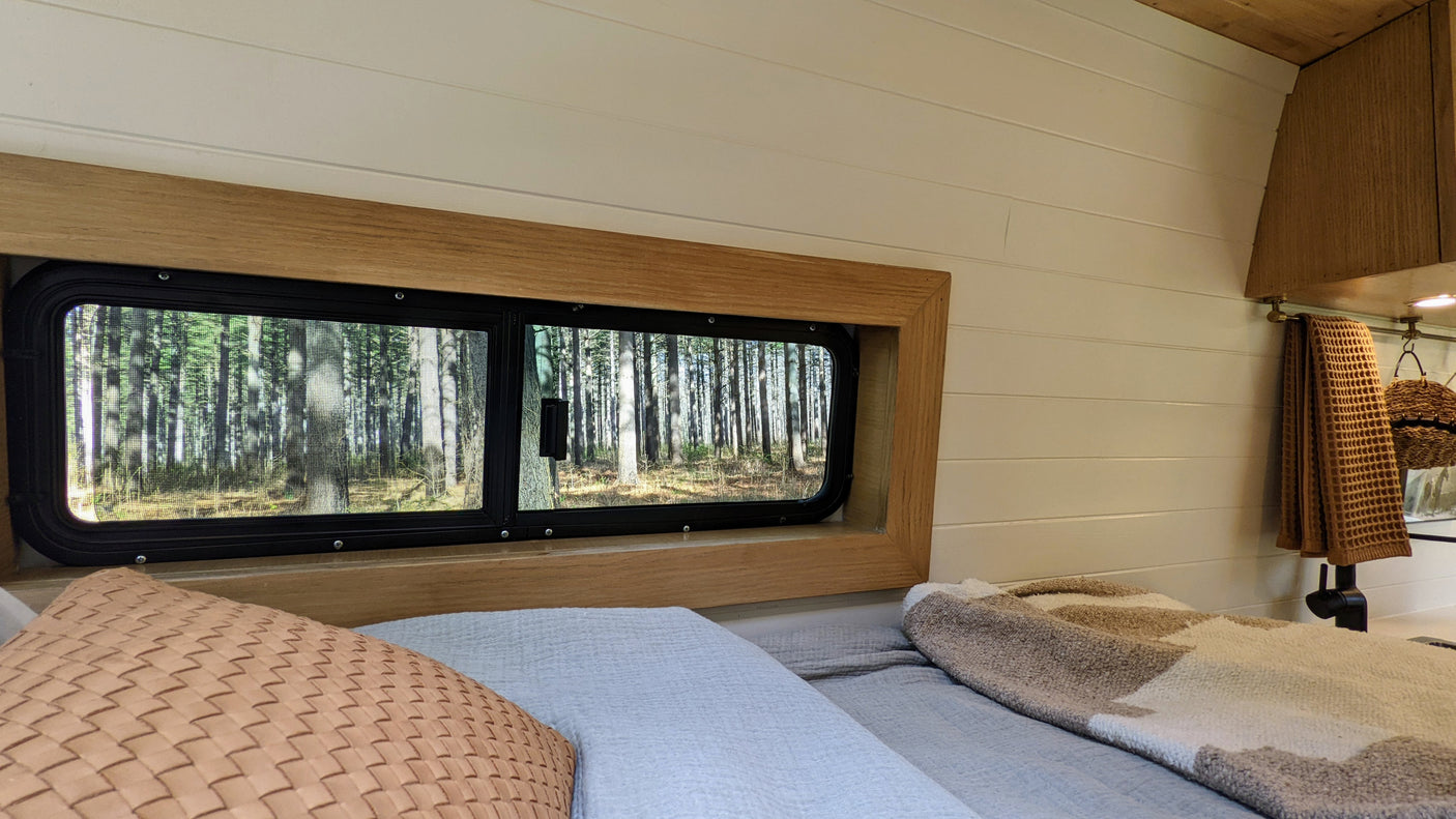 Cozy rv rental bedroom: a bed with pillows faces a window with a sunlit forest view. White shiplap walls & wood trim.