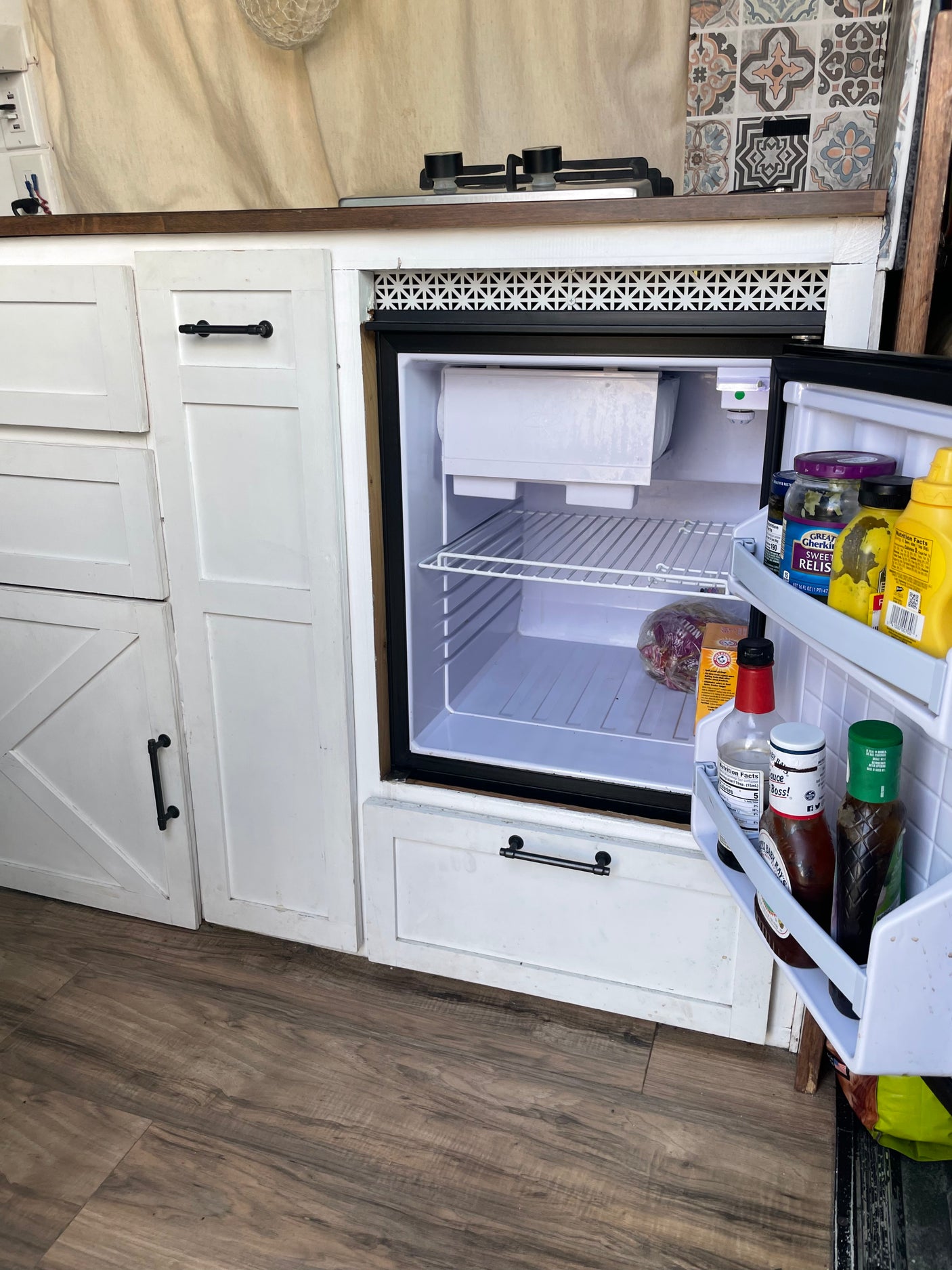 Inside this RV rental: a compact kitchen with an open mini-fridge, small stovetop, and white cabinets. Ready for your adventure!