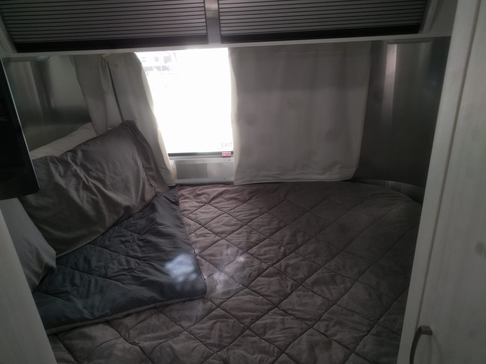 Cozy RV rental bedroom with a plush quilted bed, pillows, and a bright window featuring an "EXIT" sign.