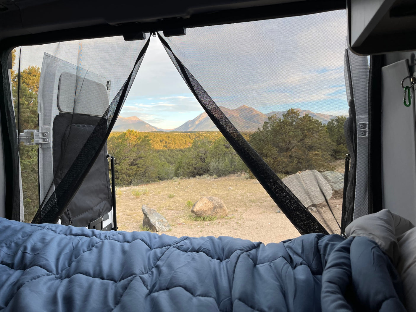 with a stunning mountain sunset. Great for remote adventures!|Cozy RV rental bed view through bug screens to majestic mountains and serene forest. Perfect for outdoor escape.|White Winnebago Solis RV rental parked on a rugged mountain road under cloudy skies