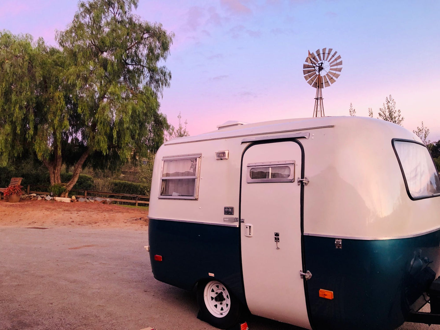 A stylish vintage RV rental, white and green, sits under a vibrant pink and blue sky next to a large tree and a rooftop windmill.