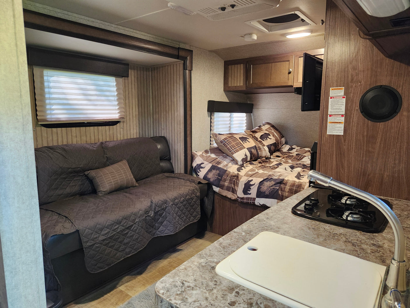 Cozy RV rental interior: dark gray couch, patterned bed, kitchen with stove and sink. Wood accents and windows.