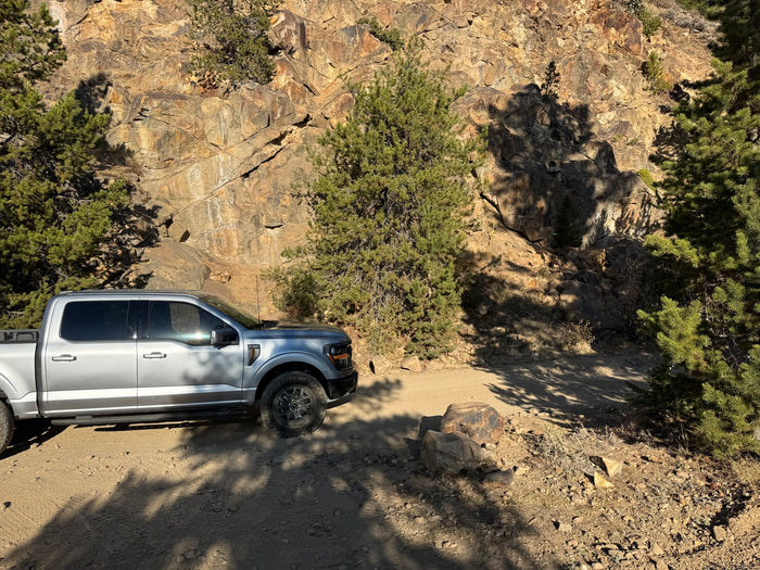 Silver pickup truck on a rugged dirt road alongside a rocky cliff & pine trees. An ideal setting for an RV rental getaway.