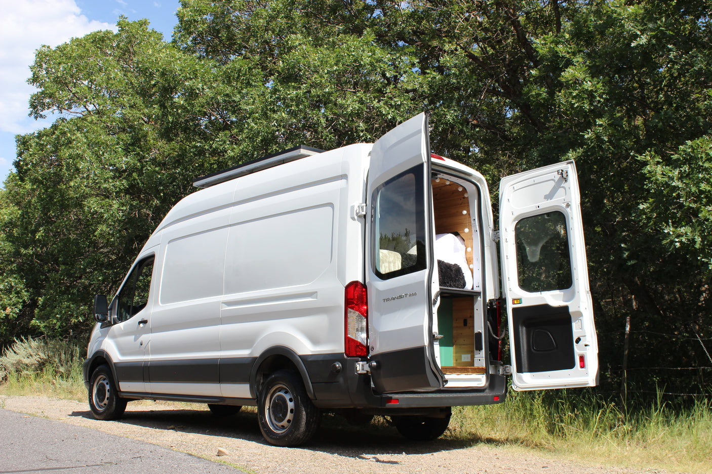 This white Ford Transit camper van, ideal for RV rental, is parked outdoors with its rear doors open, revealing a cozy wooden interior.