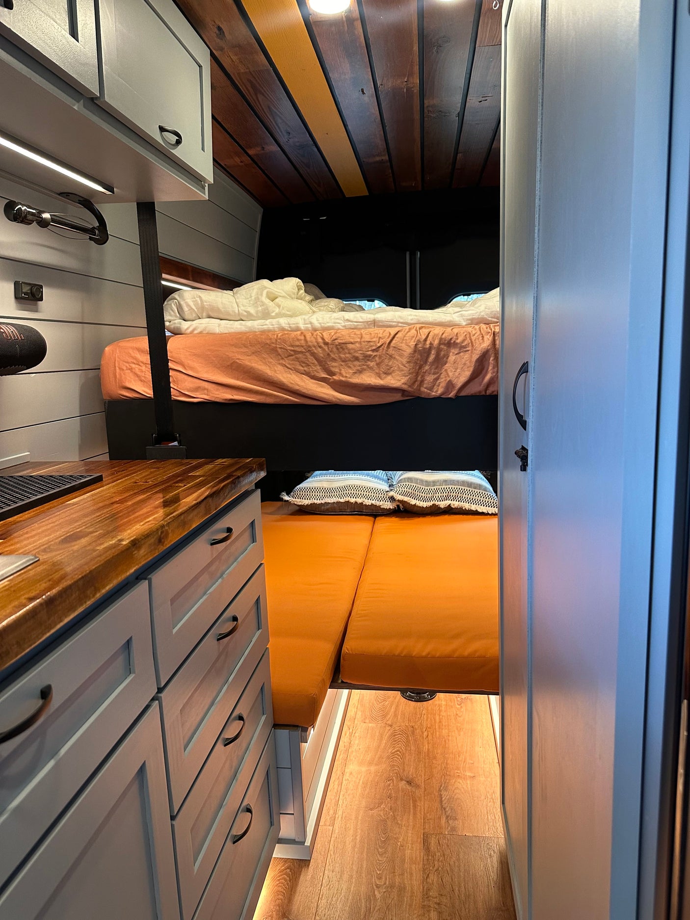 RV rental interior with light grey cabinets, wood counters, cozy upper bed, and lower orange cushion seating.
