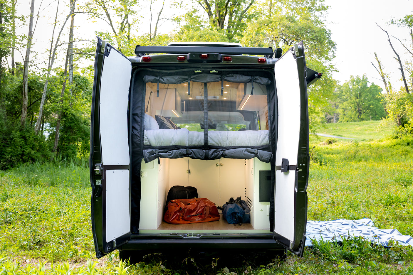 This RV rental's rear doors open to reveal a comfortable bed, storage, and lush green surroundings for your adventure.