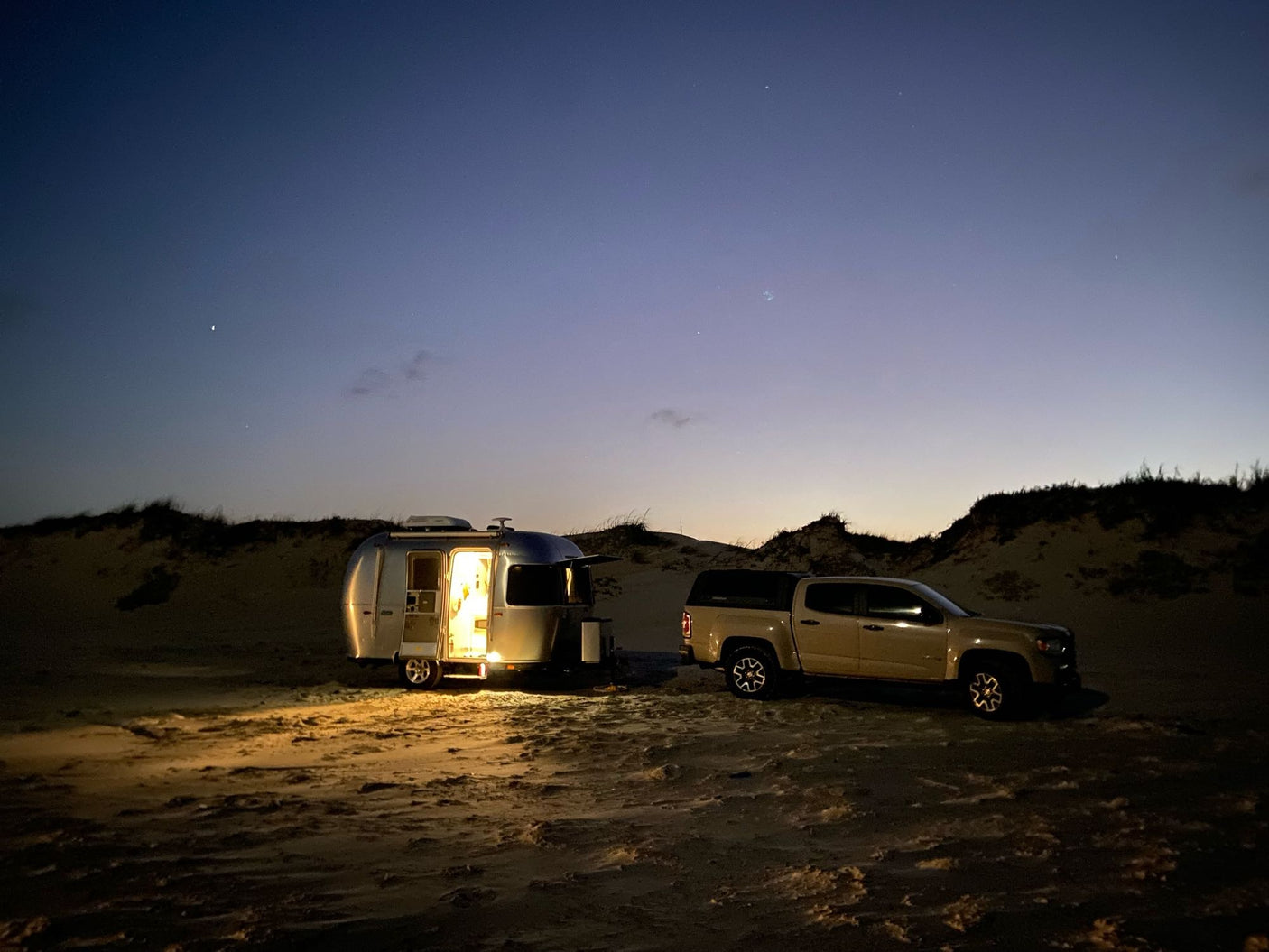 arid landscape.|Lighted RV rental and truck parked for beach camping on sand dunes under a starry twilight sky.|RV rental journey begins! An Airstream trailer next to Arizona's welcome sign on a sunny desert roadside.|Desert adventure from an RV rental! Enjoy scenic views of saguaro cacti