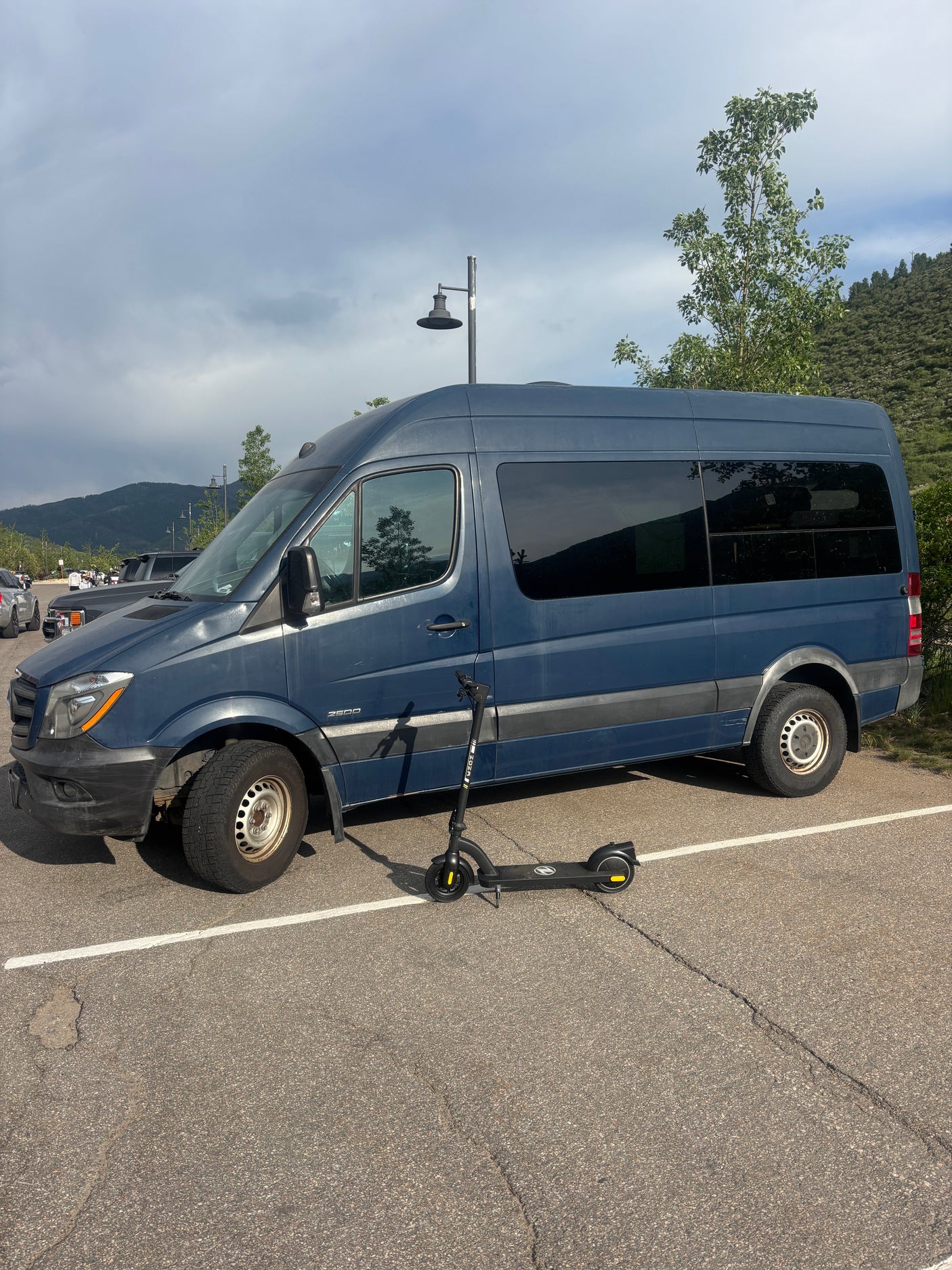 Blue Mercedes Sprinter camper van, suitable for RV rental, with scooter in a mountain lot under cloudy skies.