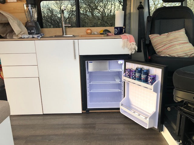Inside your RV rental: a compact kitchen featuring a bright, open fridge, sink, and clean white cabinets.