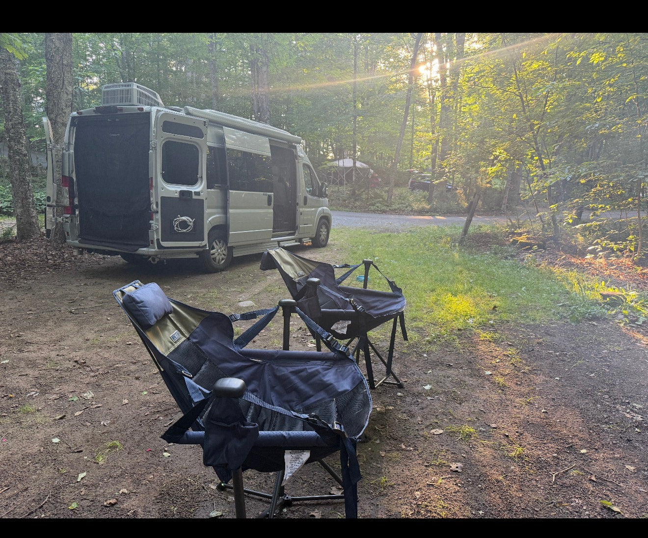 An rv rental camper van at a serene campsite with two camping chairs. Sunbeams filter through the trees. — photo 1