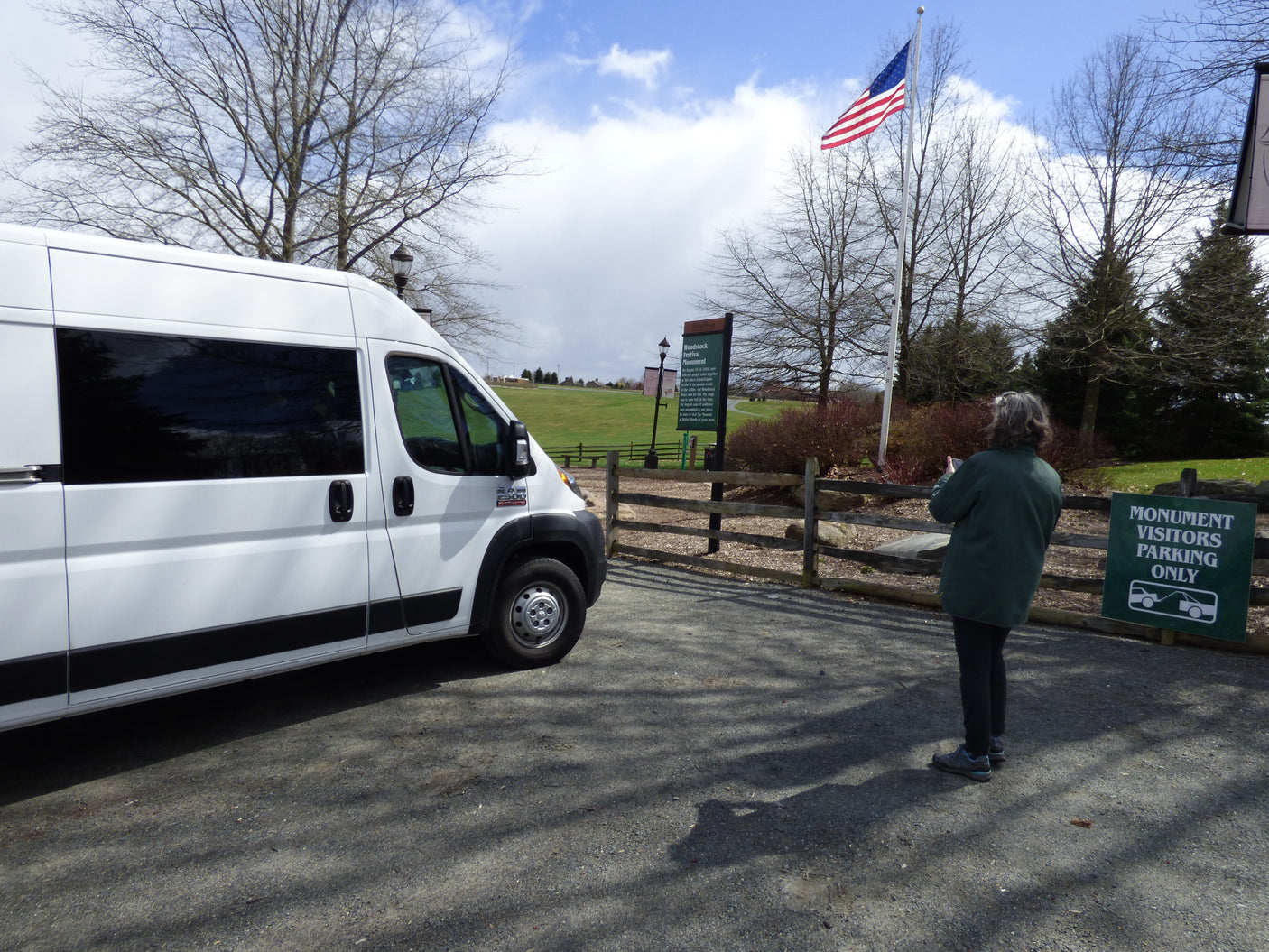 An RV rental-style van at the Woodstock Festival Monument, with a visitor viewing the historic grounds and American flag.