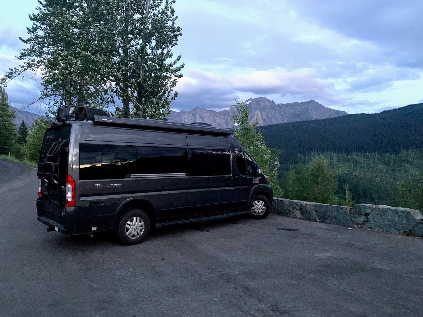 Scenic RV rental: Dark grey camper van parked on a mountain road overlooking forests & peaks under cloudy skies.