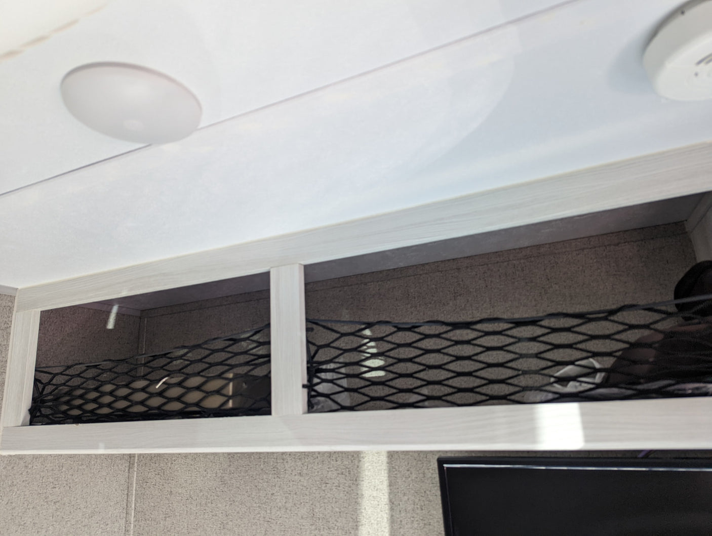 RV rental interior: overhead storage compartments with elastic netting for securing items, a ceiling light, and a smoke detector.