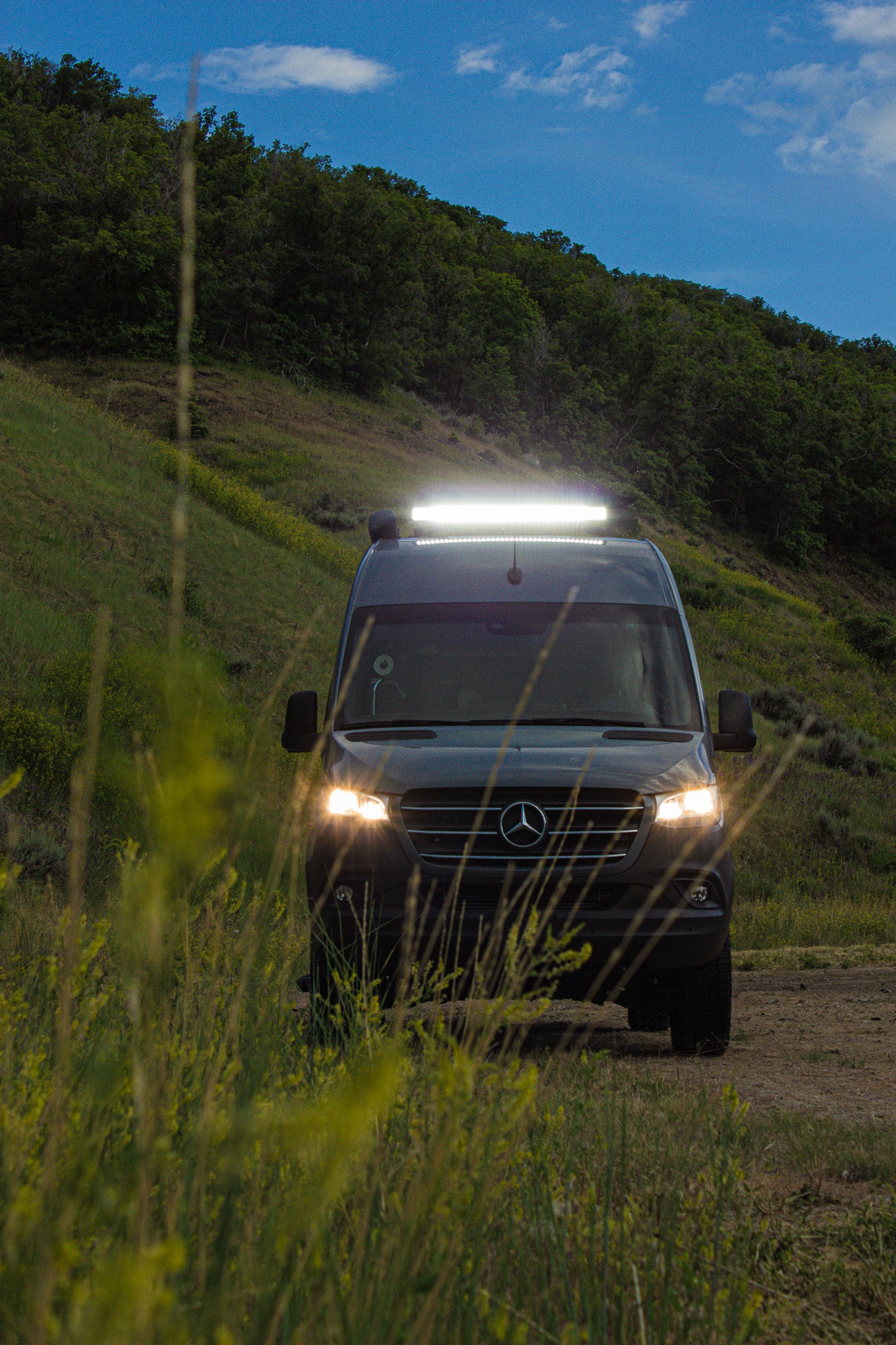 Dark Mercedes Sprinter RV rental, lights on, parked on a dirt path through tall grass & hills at dusk.