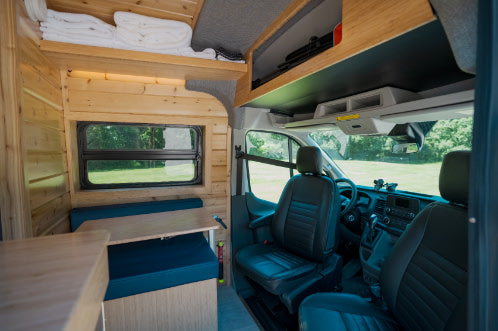 Interior of a cozy RV rental camper van with wooden walls, a blue dinette, overhead storage, and access to the front cabin.