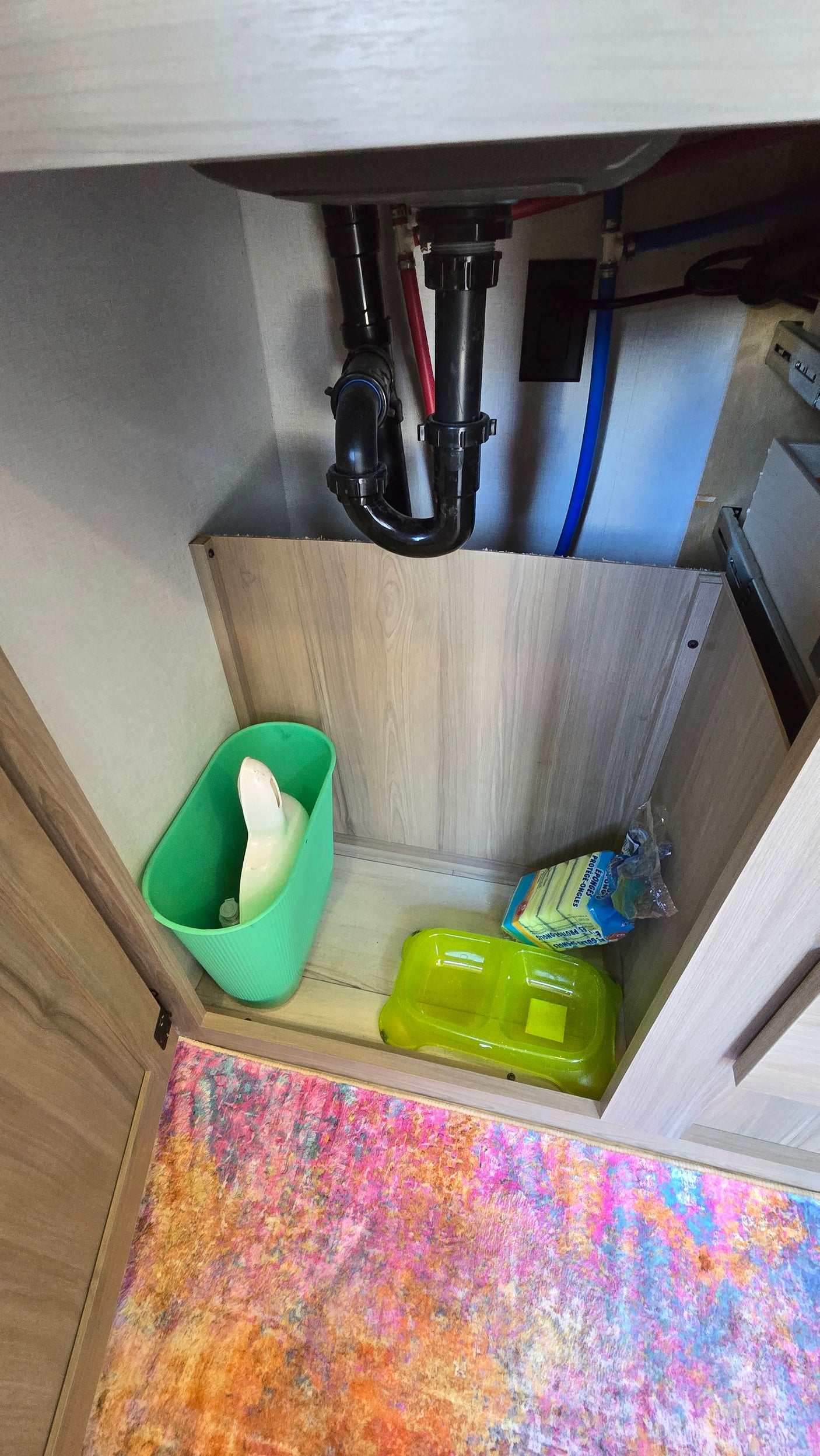 RV rental under-sink storage area, showing plumbing, a green bin, cleaning sponges, and a vibrant multi-colored rug.