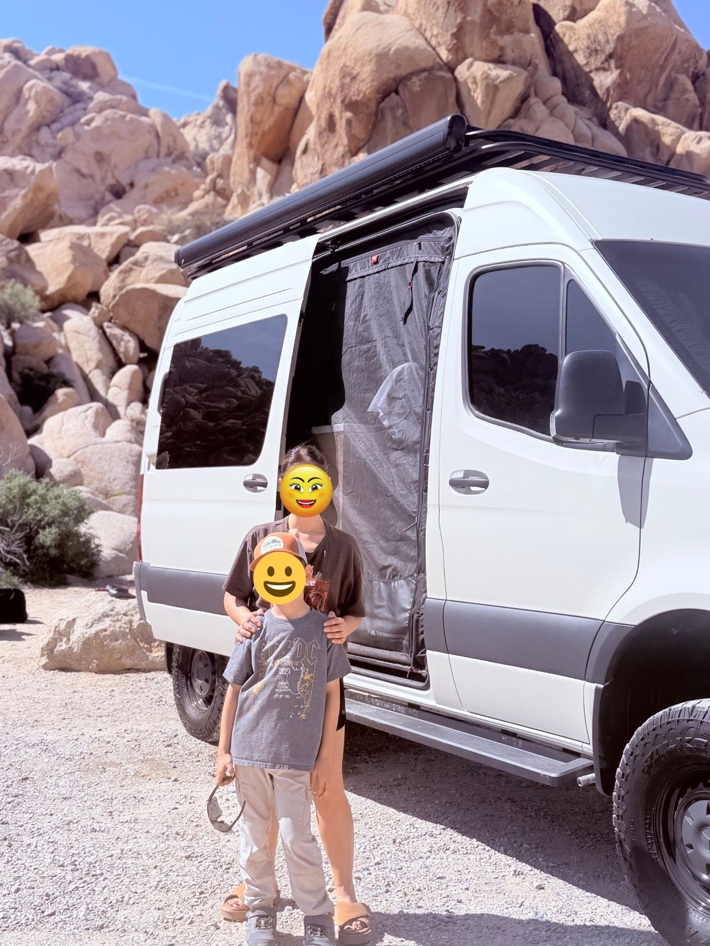Desert RV rental adventure: white camper van with awning, adult & child standing by its open door amidst large rocks.