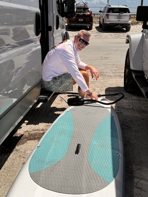 Smiling man inflates his paddleboard next to his RV rental, ready for adventure by the water.