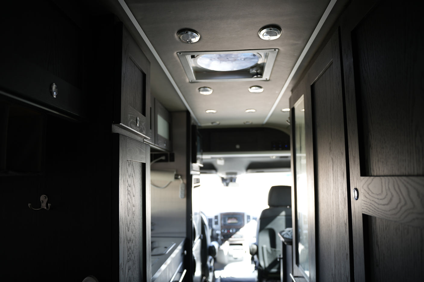 Modern RV rental interior with dark wood cabinets, ceiling lights, and the sunlit driver's area ahead.