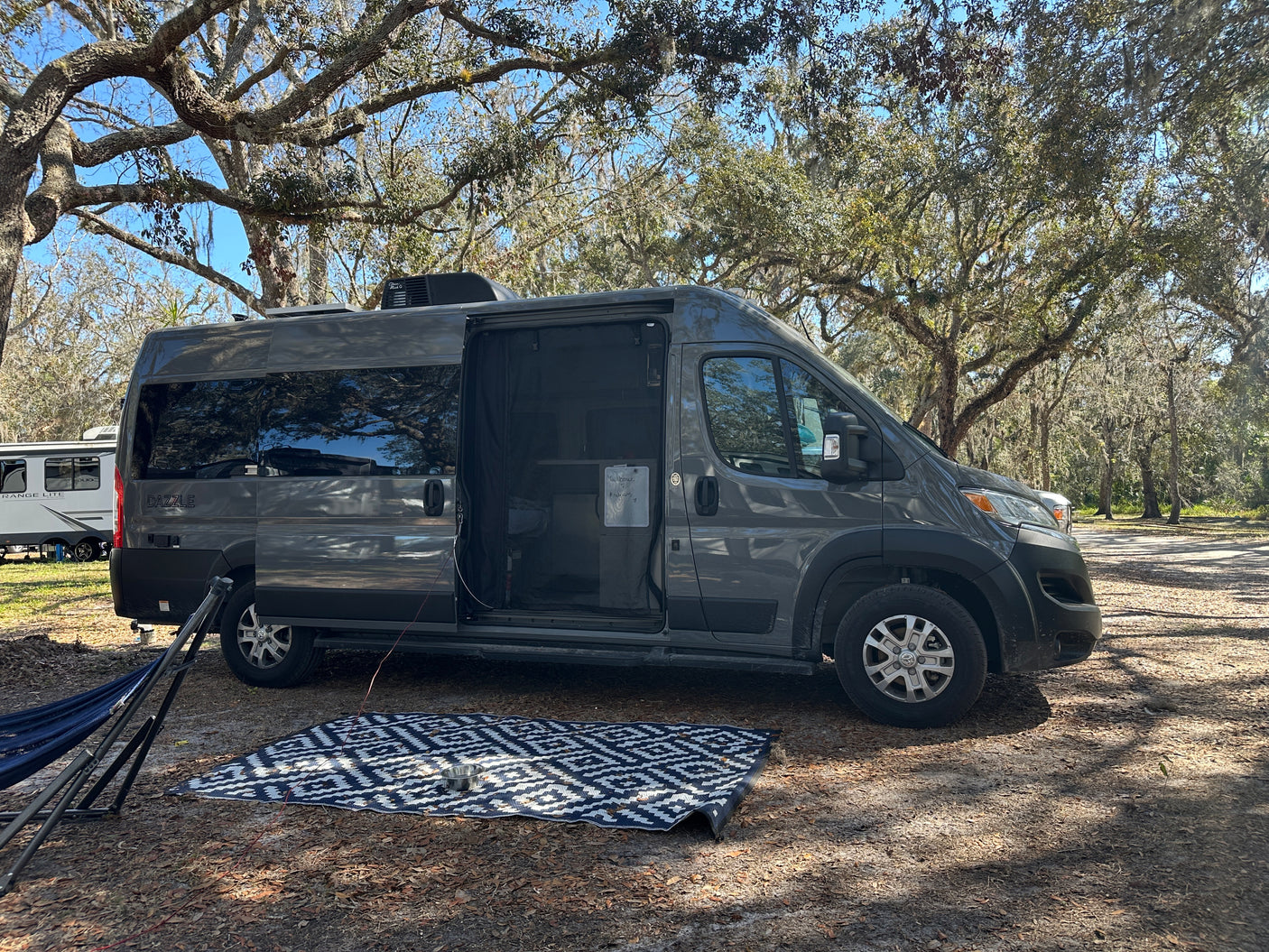 A sleek grey RV rental, with its side door open to an outdoor mat & hammock, is perfectly set in a serene, tree-filled campsite.