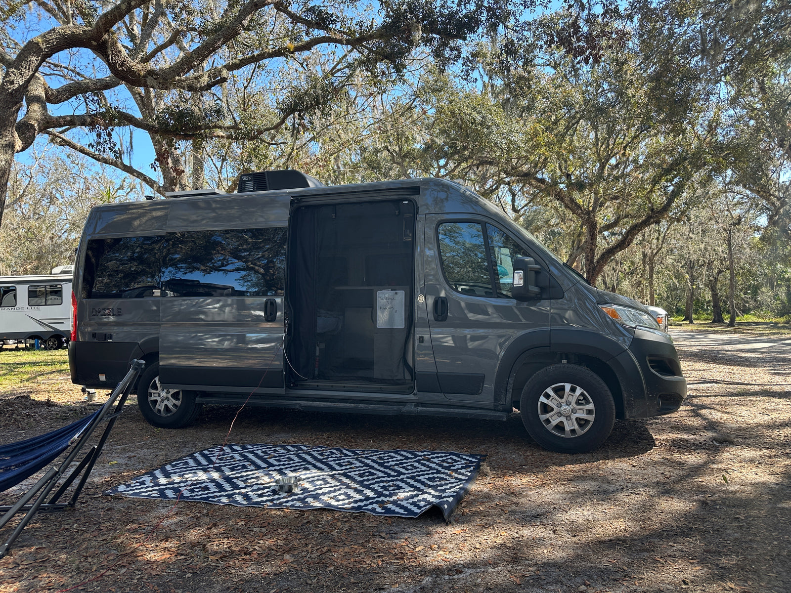 A sleek grey RV rental, with its side door open to an outdoor mat & hammock, is perfectly set in a serene, tree-filled campsite.