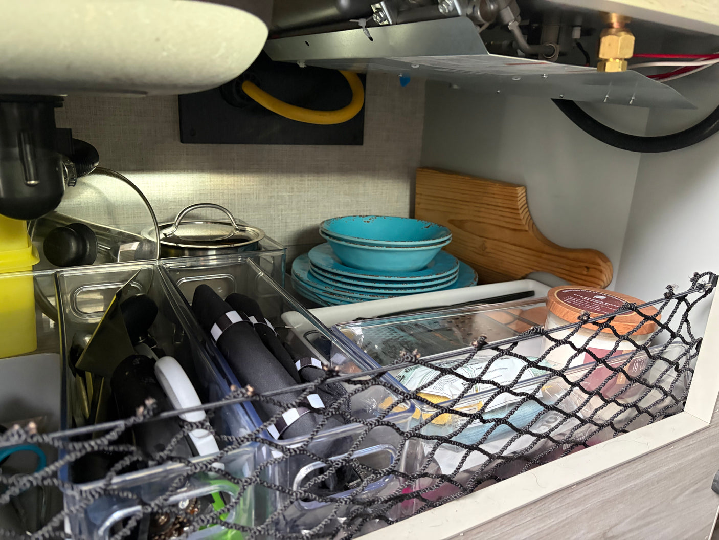 Compact kitchen storage under the sink in an RV rental, with stacked blue dishes, utensils, and organizers.