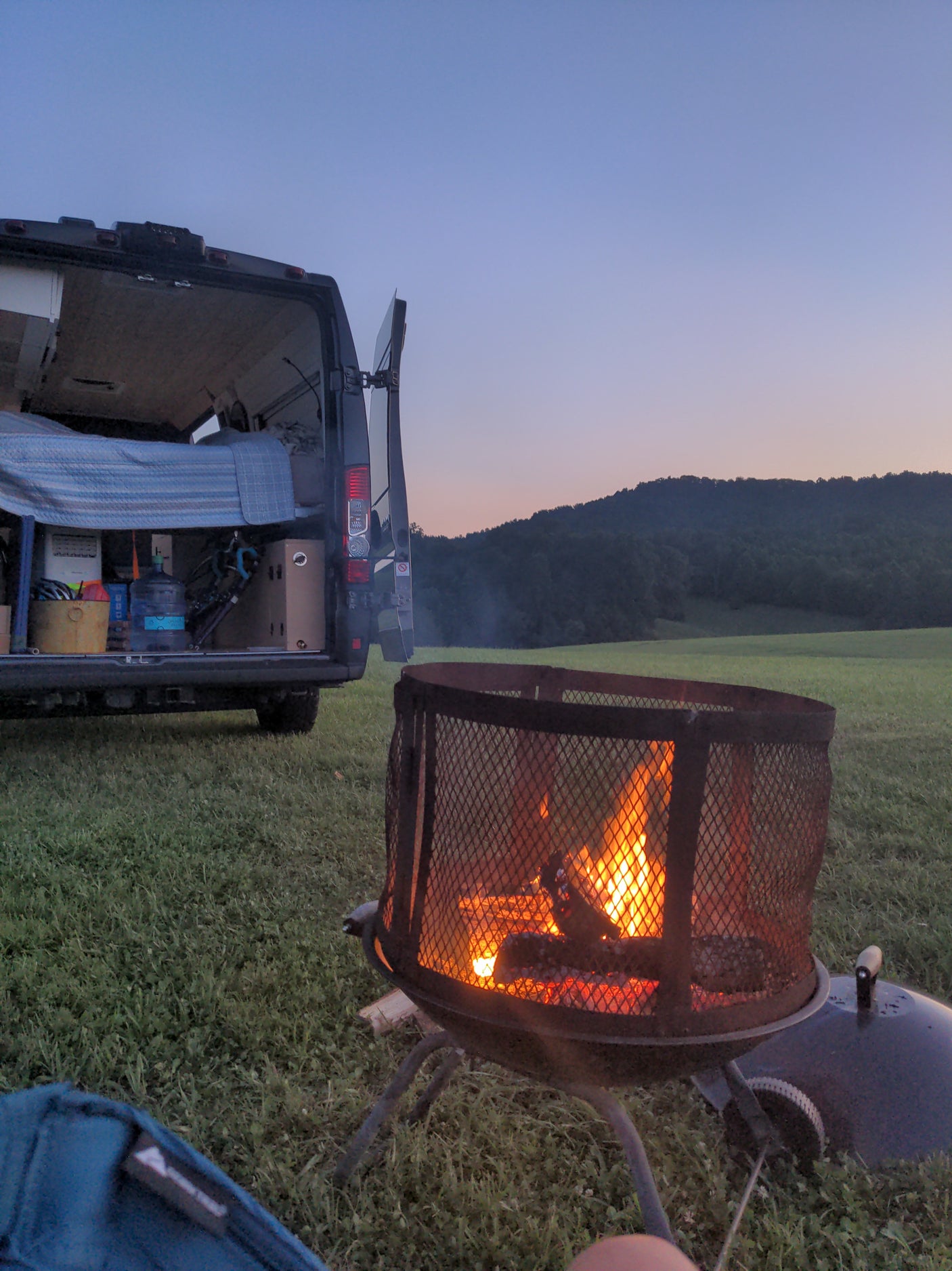 Twilight camping with an rv rental. A cozy campfire glows before a camper van in a grassy field, surrounded by hills.