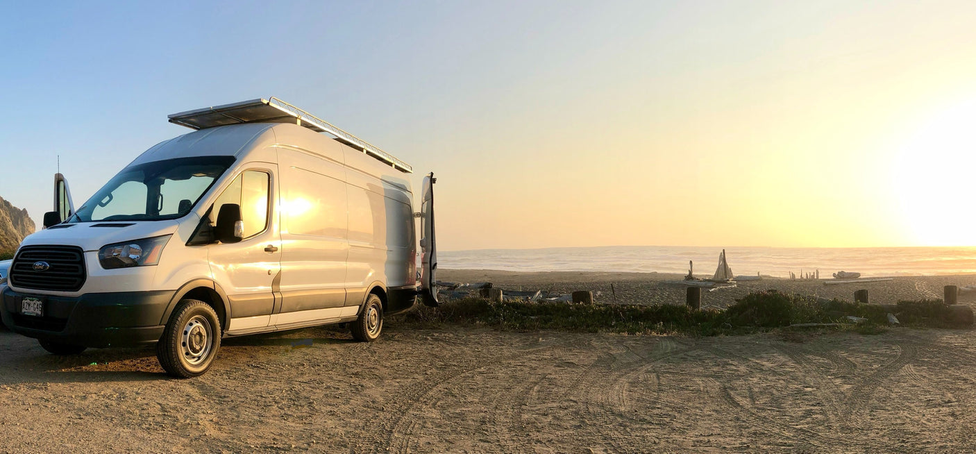 Enjoying beach camping in a white Ford Transit RV rental, parked on sand with solar panels, overlooking the ocean at sunset.