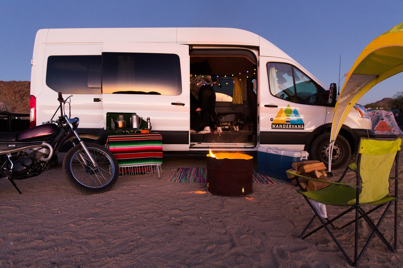 Experience desert van life with an RV rental! A Wandervans camper, campfire, & dirt bike set for adventure at dusk.