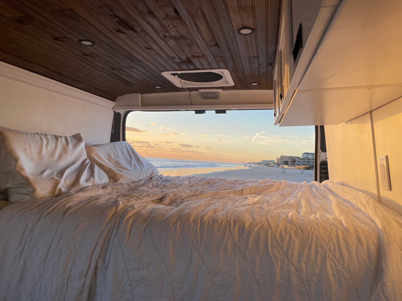Cozy RV rental bed with pillows, offering stunning ocean views of a tranquil beach, waves, and a vibrant sunset sky.