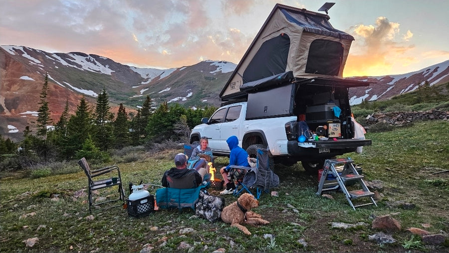 Scenic mountain camping adventure with an RV rental. A group with their dog enjoys a campfire at sunset.