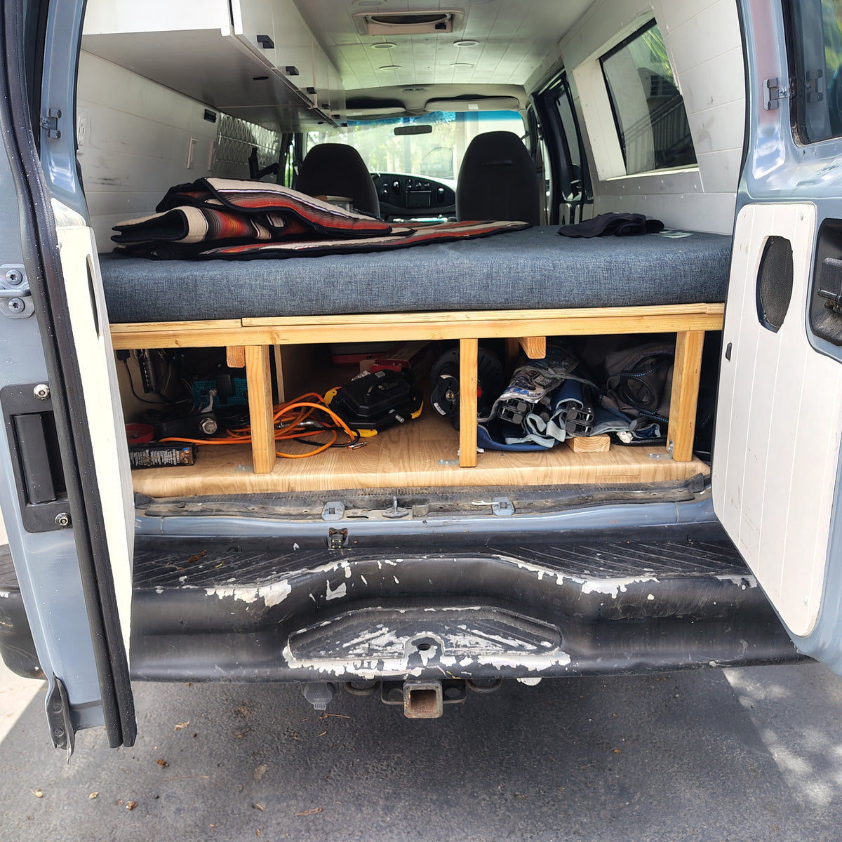 RV rental camper van interior: A blue bed cushion on a wooden platform with tools and bags stored below, seen through open rear doors. — photo 4
