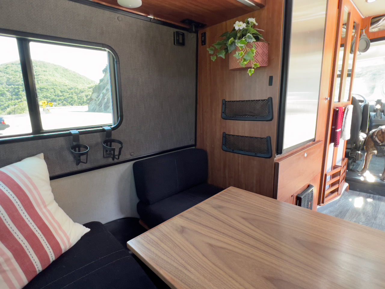 Inside a comfy RV rental: dining table, window with scenic mountain road, seating area, and a dog visible in the front.