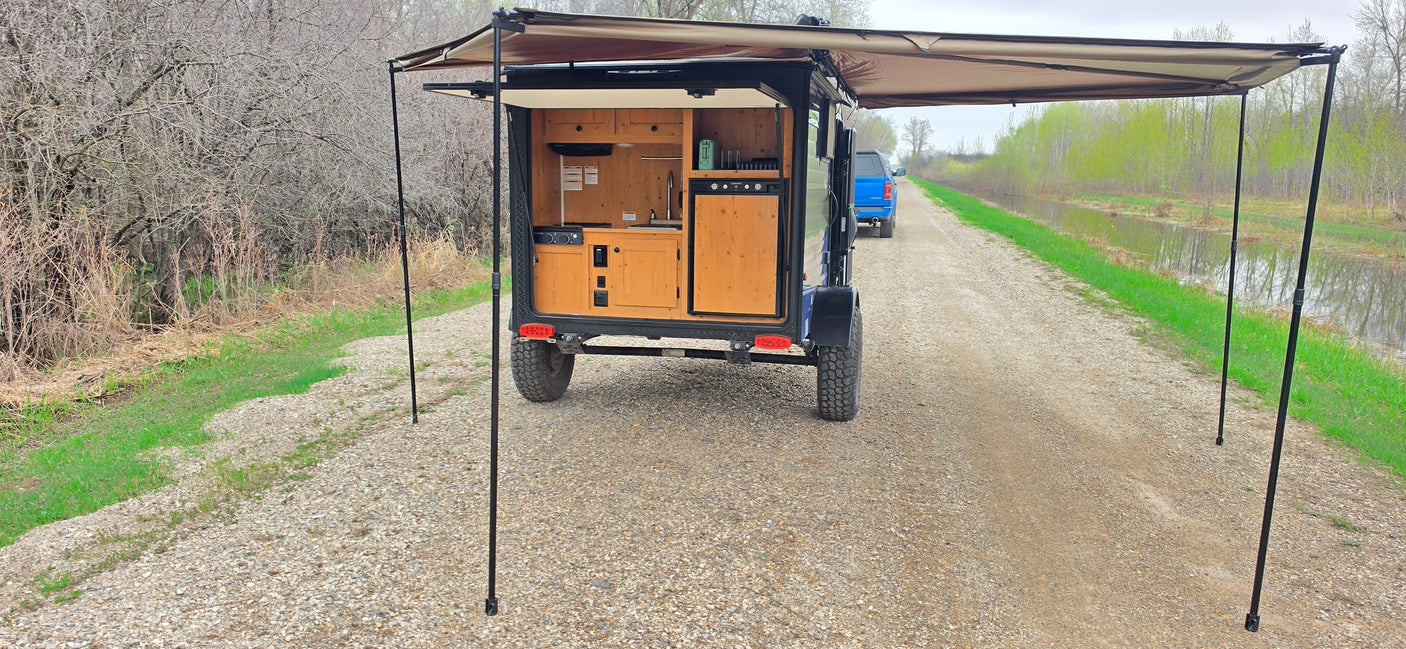 RV rental: Small camper trailer with outdoor kitchen & awning, parked on a gravel road next to a canal. Ready for adventure.