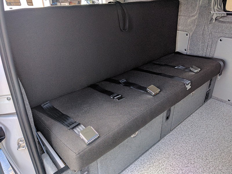 RV rental: Versatile camper van interior showing a dark grey bench seat with three lap seatbelts & built-in storage.