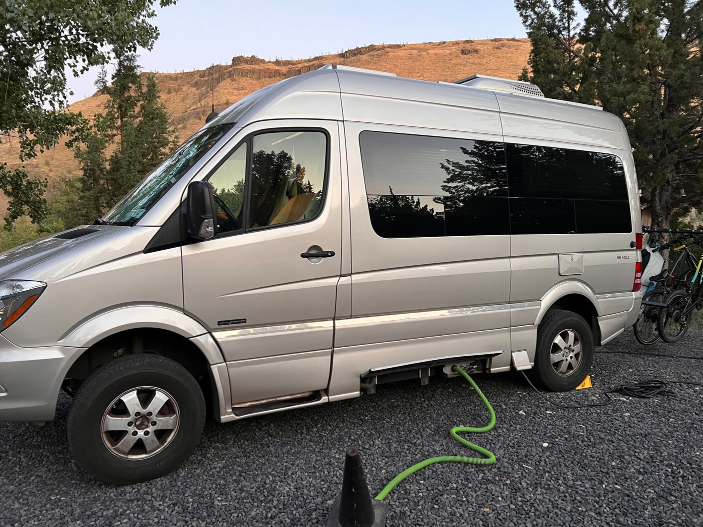 Silver Roadtrek RV rental (camper van) at a gravel campsite with hookups, bikes, and mountain views.