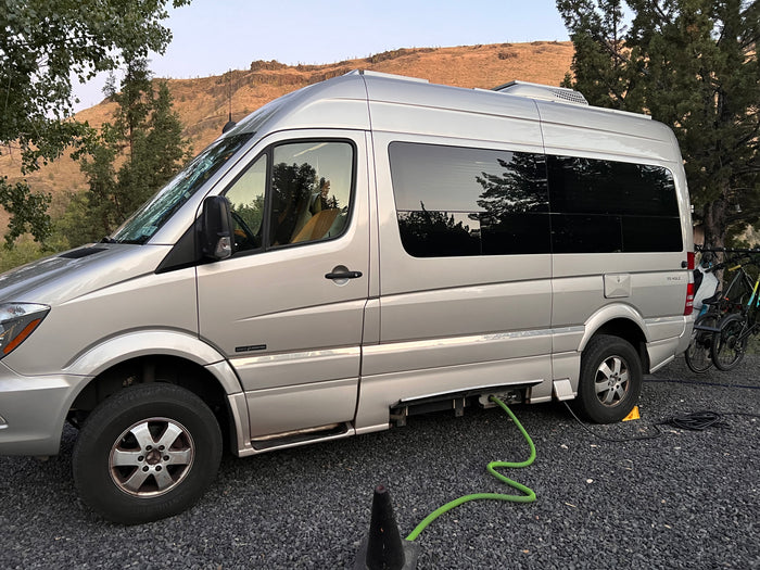 Silver Roadtrek RV rental (camper van) at a gravel campsite with hookups, bikes, and mountain views.