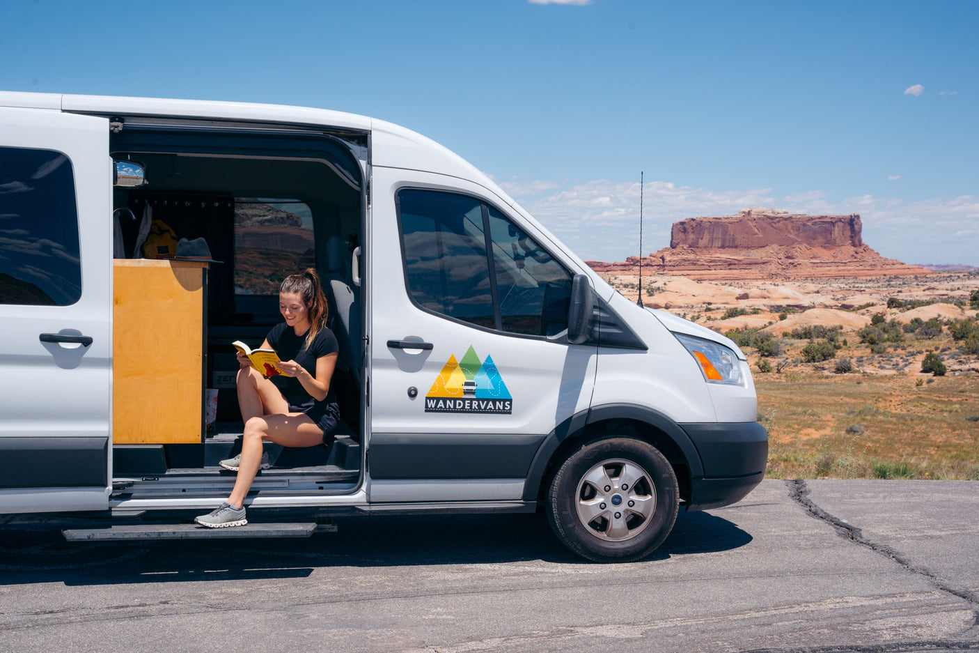 Relax in your Wandervans RV rental! A woman enjoys a book from her camper van amidst stunning desert mesas.
