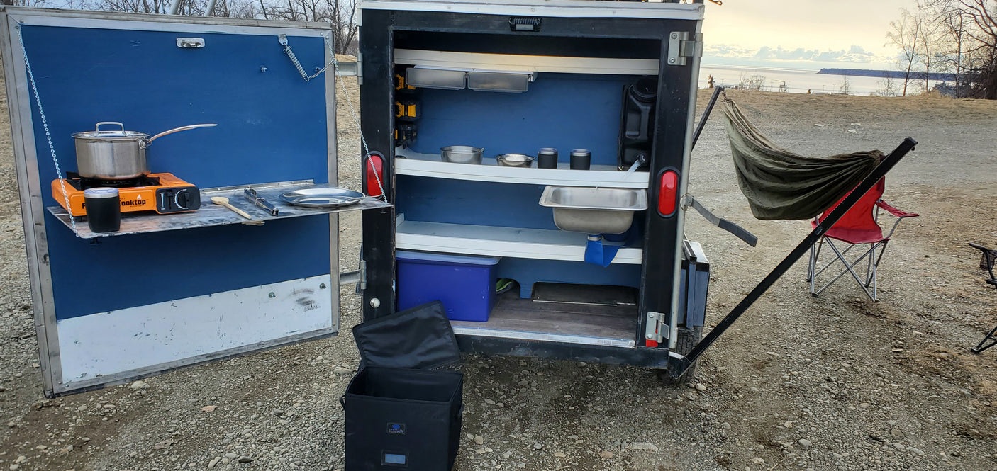 Compact RV rental with an innovative outdoor kitchen featuring a portable stove, pot, sink, and shelves for roadside cooking by the water.