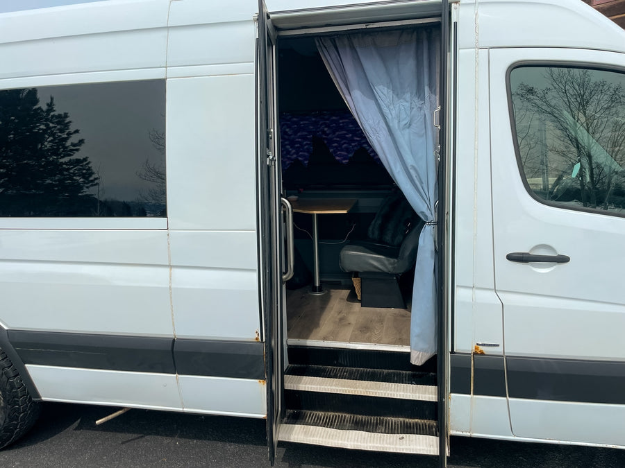 This white RV rental reveals its open side door, showcasing a cozy interior with a table and seating, ready for your next van life adventure.