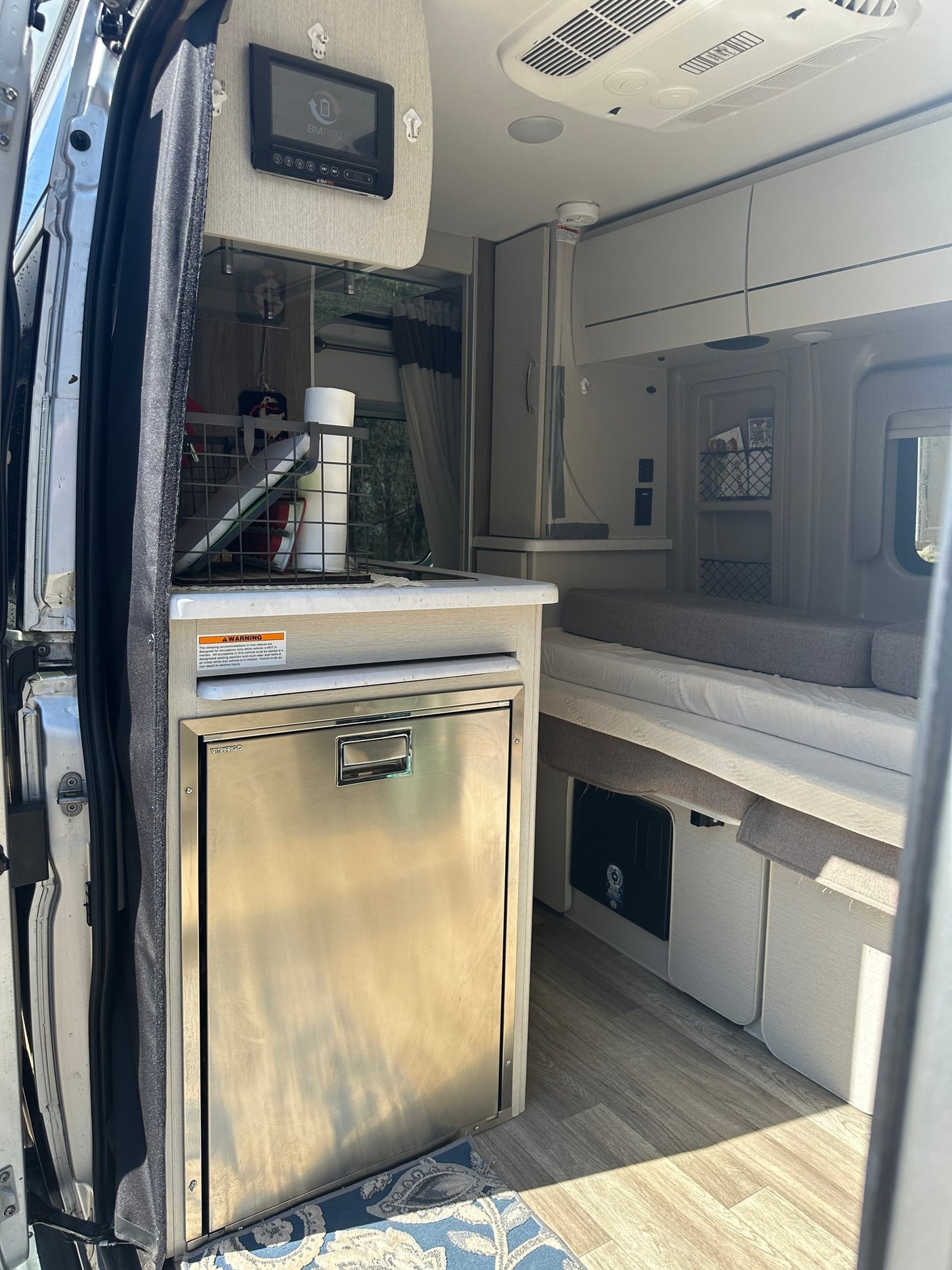 Inside a modern rv rental: stainless fridge, counter with supplies, and comfortable bed/seating. Clean, well-lit interior.