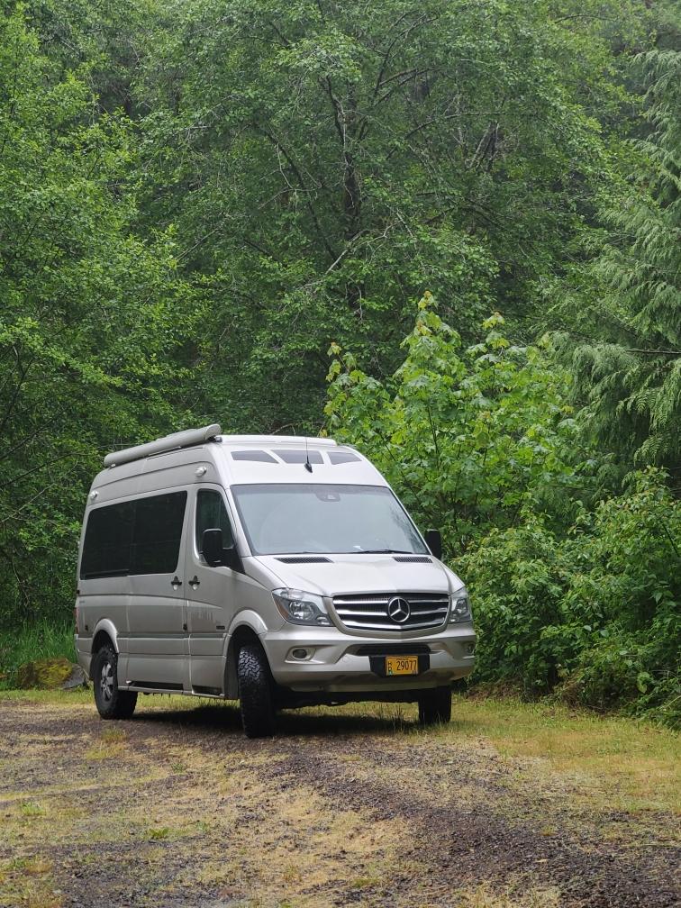 parked on a dirt road amidst lush green forest.|Silver Roadtrek RV rental (camper van) at a gravel campsite with hookups — photo 3
